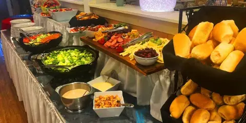 A catering buffet table featuring a salad, charcuterie board, cheese, and baskets of bread rolls.