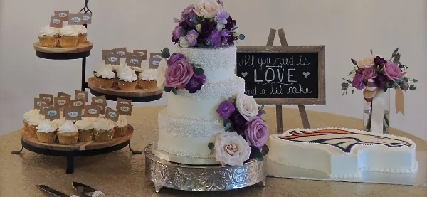 A three-tiered white wedding cake with purple and white flowers, displayed next to a tiered stand of cupcakes and a heart-shaped cake.