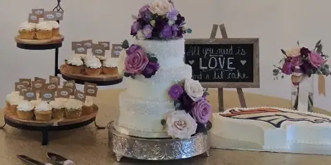 A three-tiered white wedding cake with purple and white flowers, displayed next to a tiered stand of cupcakes and a heart-shaped cake.