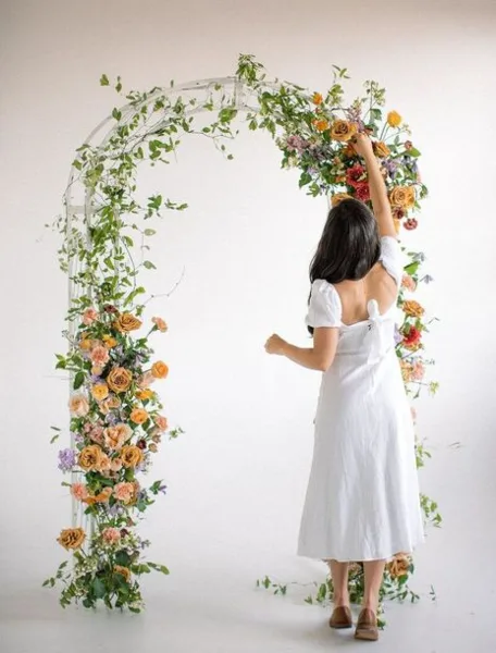 A woman in a white dress arranging a large floral arch with roses and greenery.