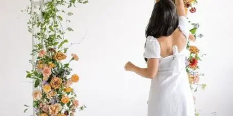 A woman in a white dress arranging a large floral arch with roses and greenery.