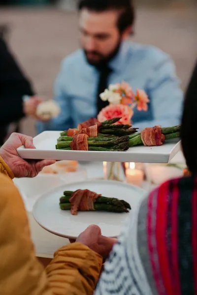 A server holding a white platter of prosciutto-wrapped asparagus at an outdoor event.