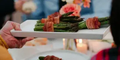 A server holding a white platter of prosciutto-wrapped asparagus at an outdoor event.
