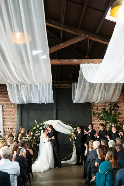 Wedding couple kissing during a ceremony in an industrial venue with exposed brick walls and white drapery.
