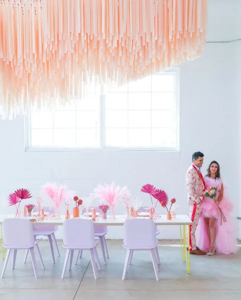 A bride and groom standing in a bright, modern warehouse event space with a large pink hanging installation and a decorated table.