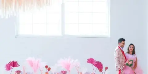 A bride and groom standing in a bright, modern warehouse event space with a large pink hanging installation and a decorated table.