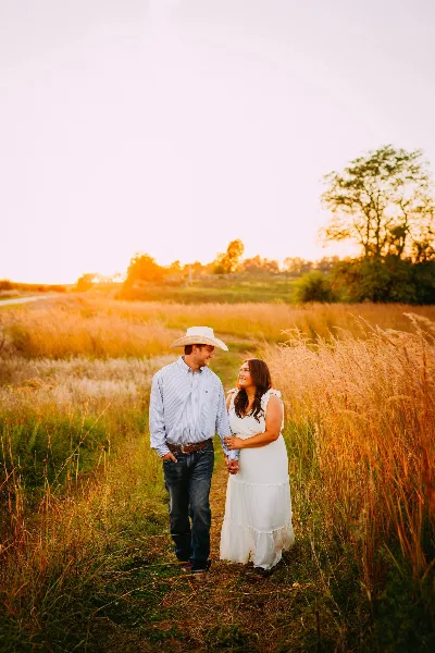 A couple holding hands in a grassy field at sunset, with the man wearing a cowboy hat and the woman in a white dress.