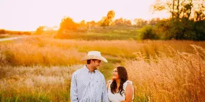 A couple holding hands in a grassy field at sunset, with the man wearing a cowboy hat and the woman in a white dress.