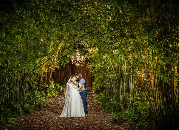 A bride and groom embracing in a garden path lined with bamboo and ferns, with a chandelier hanging above a rustic wooden structure in the background.