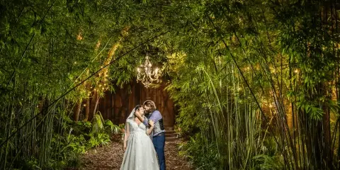 A bride and groom embracing in a garden path lined with bamboo and ferns, with a chandelier hanging above a rustic wooden structure in the background.