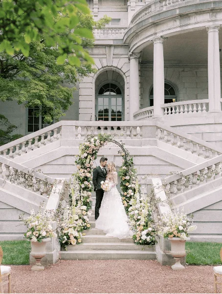 A bride and groom standing on the grand stone staircase of a historic estate, framed by a floral arch and large flower urns.