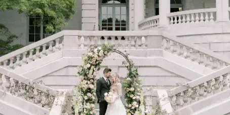 A bride and groom standing on the grand stone staircase of a historic estate, framed by a floral arch and large flower urns.