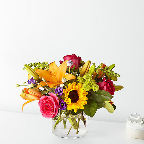 A vibrant floral arrangement featuring orange lilies, pink roses, and a sunflower in a clear glass vase.