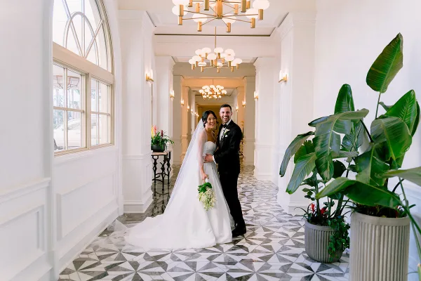A bride and groom standing together in a white hallway with geometric tile flooring and large potted plants.