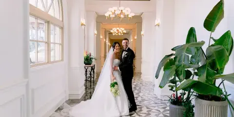 A bride and groom standing together in a white hallway with geometric tile flooring and large potted plants.