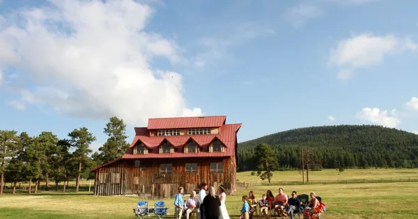 A bride and groom stand before an officiant and seated guests in a grassy field in front of a rustic wooden barn with a red roof.
