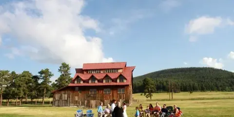 A bride and groom stand before an officiant and seated guests in a grassy field in front of a rustic wooden barn with a red roof.