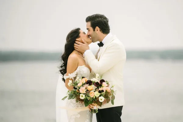 A bride and groom sharing a kiss outdoors near a body of water, with the bride holding a floral bouquet.