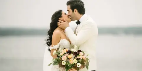 A bride and groom sharing a kiss outdoors near a body of water, with the bride holding a floral bouquet.