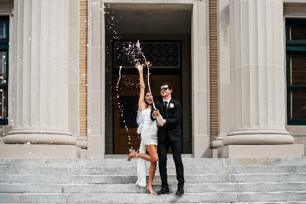 A bride and groom standing on stone steps outside a building, celebrating with a champagne bottle spraying.
