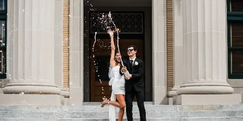 A bride and groom standing on stone steps outside a building, celebrating with a champagne bottle spraying.