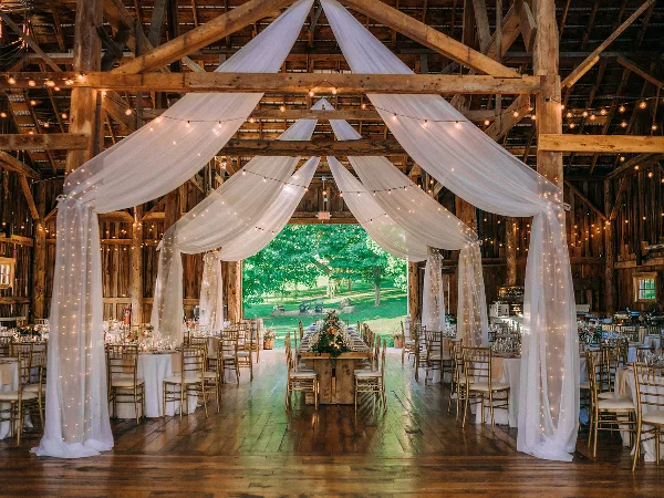 Interior of a rustic barn wedding venue featuring exposed wooden beams, white drapery, string lights, and a long dining table set for a reception.