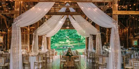 Interior of a rustic barn wedding venue featuring exposed wooden beams, white drapery, string lights, and a long dining table set for a reception.