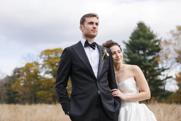 A bride and groom posing together in a field with autumn trees in the background.