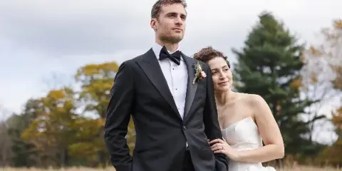 A bride and groom posing together in a field with autumn trees in the background.