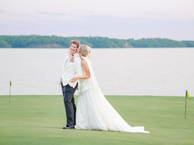 A bride and groom embracing on a grassy field near a body of water.