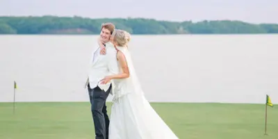 A bride and groom embracing on a grassy field near a body of water.