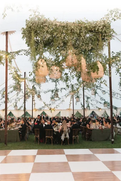 A luxury wedding reception inside a large tent featuring a massive hanging floral installation with woven pendant lights and a checkered dance floor.
