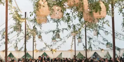 A luxury wedding reception inside a large tent featuring a massive hanging floral installation with woven pendant lights and a checkered dance floor.