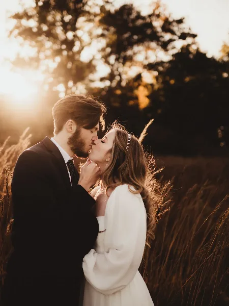 A bride and groom sharing a kiss in a sunlit field with tall grass and trees in the background.