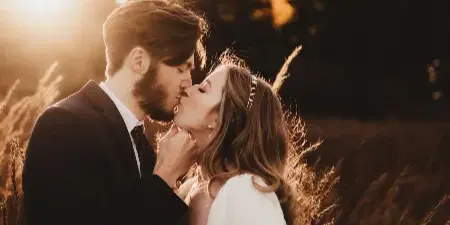A bride and groom sharing a kiss in a sunlit field with tall grass and trees in the background.