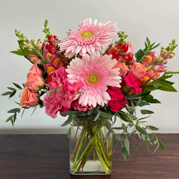 A floral arrangement with pink gerbera daisies, roses, and greenery in a clear glass vase.