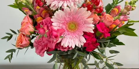 A floral arrangement with pink gerbera daisies, roses, and greenery in a clear glass vase.
