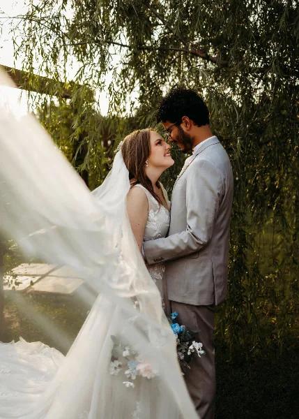 A bride and groom standing close together under a tree, with the bride's veil flowing in the foreground.