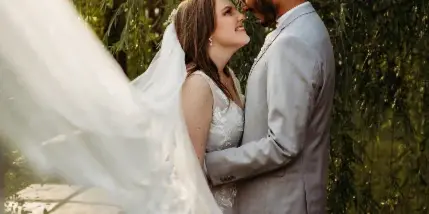 A bride and groom standing close together under a tree, with the bride's veil flowing in the foreground.
