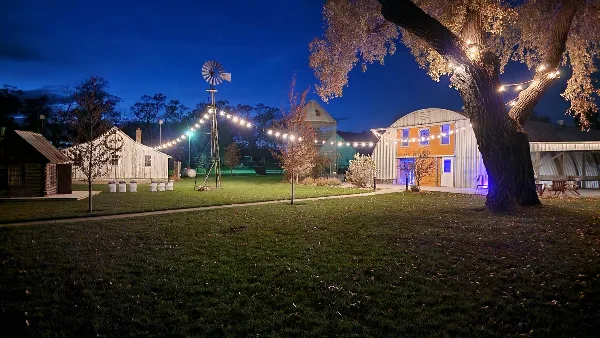 A wide shot of a rustic farm venue at night, featuring a windmill, string lights, and a large barn structure.