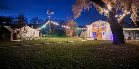A wide shot of a rustic farm venue at night, featuring a windmill, string lights, and a large barn structure.