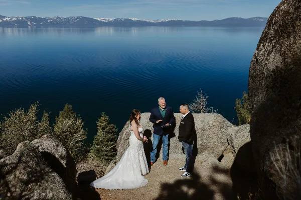 A wedding officiant performing a ceremony for a bride and groom on a rocky outcrop overlooking a large lake and mountains.