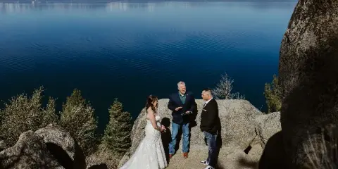 A wedding officiant performing a ceremony for a bride and groom on a rocky outcrop overlooking a large lake and mountains.