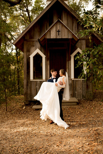 A bride and groom standing in front of a rustic wooden chapel with a cross on the roof.