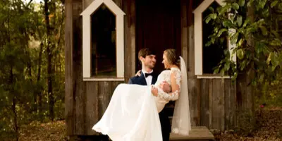 A bride and groom standing in front of a rustic wooden chapel with a cross on the roof.