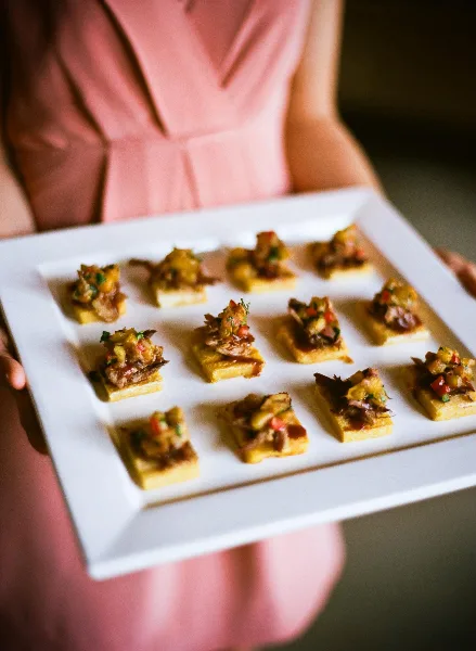 A server in a pink dress holding a white square platter filled with small, square food appetizers topped with diced vegetables and meat.