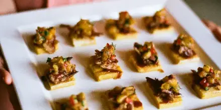 A server in a pink dress holding a white square platter filled with small, square food appetizers topped with diced vegetables and meat.