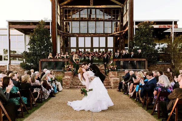 A bride and groom sharing a kiss during an outdoor wedding ceremony in front of a large, multi-story building with industrial-style windows and brick pillars, surrounded by seated guests.