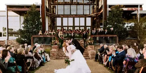 A bride and groom sharing a kiss during an outdoor wedding ceremony in front of a large, multi-story building with industrial-style windows and brick pillars, surrounded by seated guests.