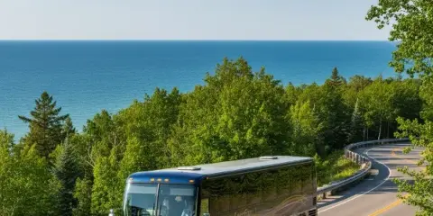 A dark blue luxury motorcoach bus driving on a scenic coastal highway with a large body of water in the background.